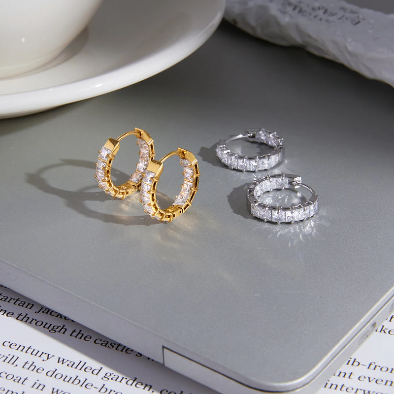 Two pairs of square crystal huggie hoops on a grey surface