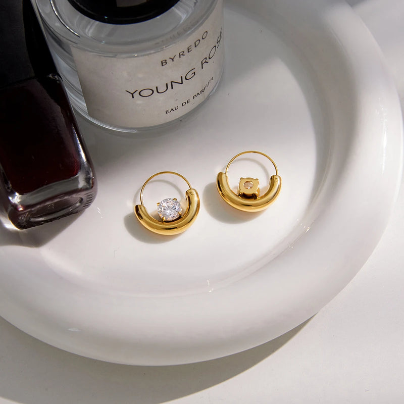 Pair of crescent crystal hoops on a white dish beside a bottle