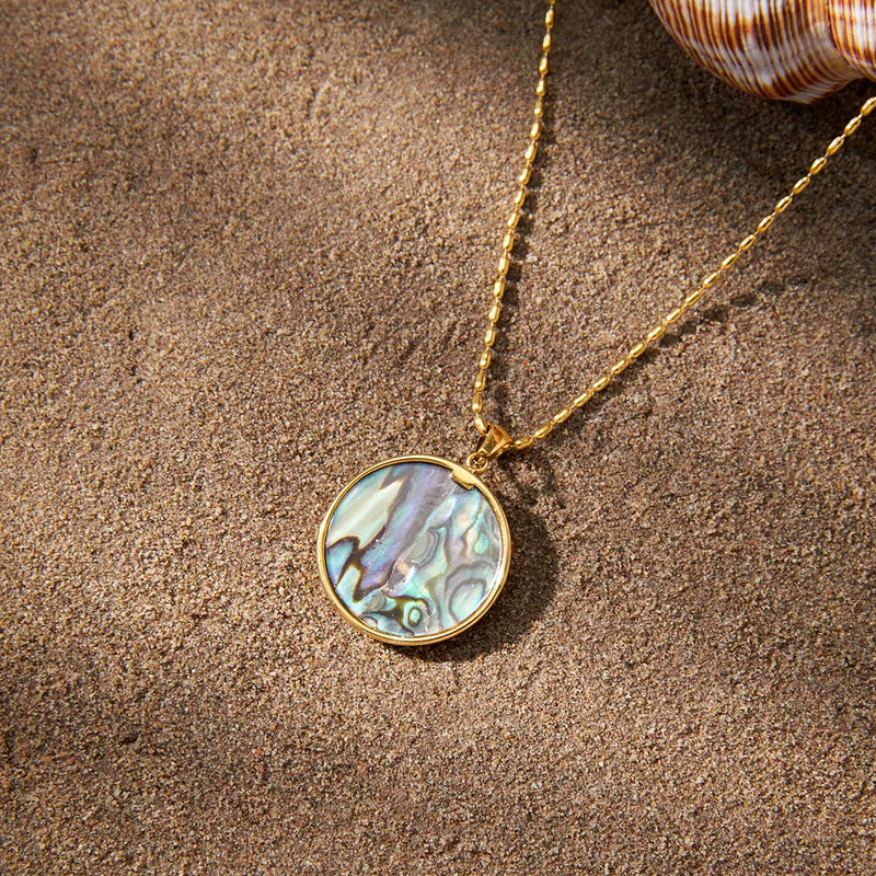 Round iridescent shell pendant on a chain, laid on sand